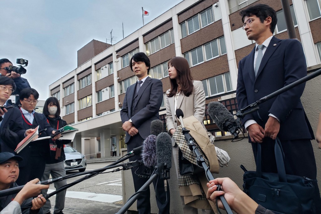 Photo by NATSUKO FUKUE / AFP.  Riho Fukuyama (C) and her husband Yoshiki Fukuyama (C-L) speak to the media with their lawyer (R) outside the Toyama District Court in Toyama city on October 21, 2025.