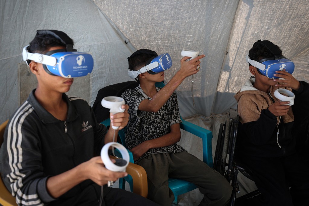 Photo by EYAD BABA / AFP  Palestinian children wearing goggles and holding a joy stick experience virtual reality as a medical technology support team launches an initiative in the city of Al-Zawayda, in the central Gaza Strip on November 30, 2025