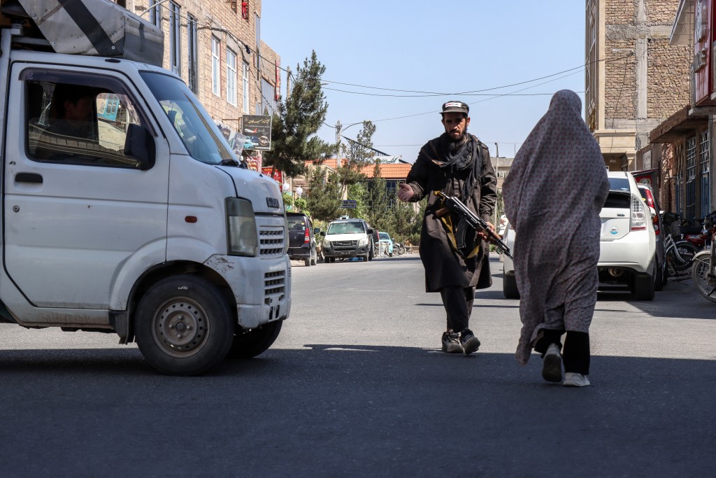 Photo by MOHSEN KARIMI / AFP  / An Afghan burqa-clad woman walks past a Taliban security personnel standing guard outside a mosque in Shahrak-e-Almahdi, Jebrail district of Herat Province on June 11, 2026.