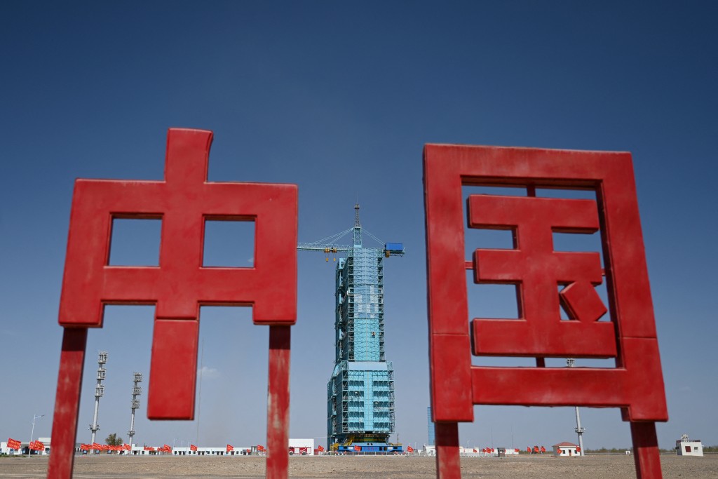 Photo by GREG BAKER / AFP  A Long March-2F carrier rocket, carrying the Shenzhou-18 spacecraft, is seen encased in a shield on the launch pad, behind a sign featuring the Chinese characters for “China”, a day before the launch of the mission, at the Jiuquan Satellite Launch Centre in the Gobi desert in northwest China on April 24, 2024.