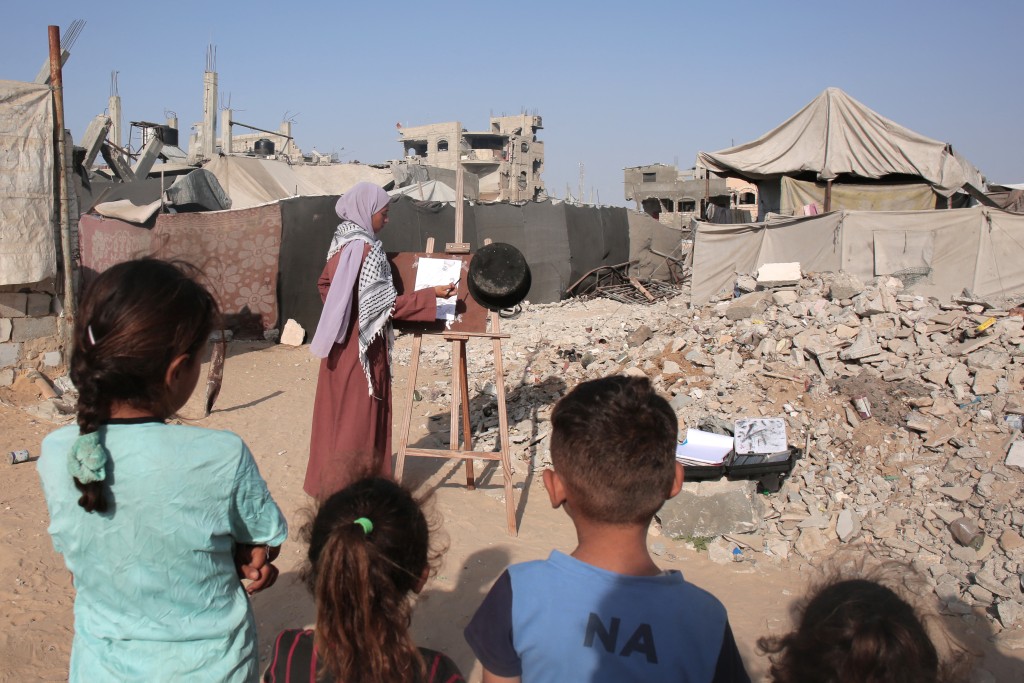 Displaced Palestinian children watch Raghda Al-Eid, a displaced 23-year-old Palestinian artist, paint using soot from the bottom of pot at a camp for the displaced in the Mawasi area of Khan Yunis on August 19, 2025. Gaza's civil defence agency reported that 45 people were killed on August 19 by Israeli strikes and fire across the territory. (Photo by AFP)