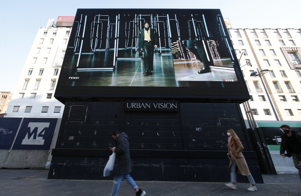 People walk past a giant screen streaming a Fendi fashion live show during the Milan's fashion week in Milan, Italy, Friday, January 15, 2021. People walk past a giant screen streaming a Fendi fashion live show during the Milan's fashion week in Milan, Italy, Friday, January 15, 2021.