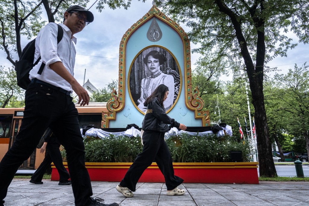 Photo by CHANAKARN LAOSARAKHAM / AFP  People, some dressed in black to mourn the death of Thailand's former queen Sirikit, walk past her portrait displayed in Bangkok on October 30, 2025.
