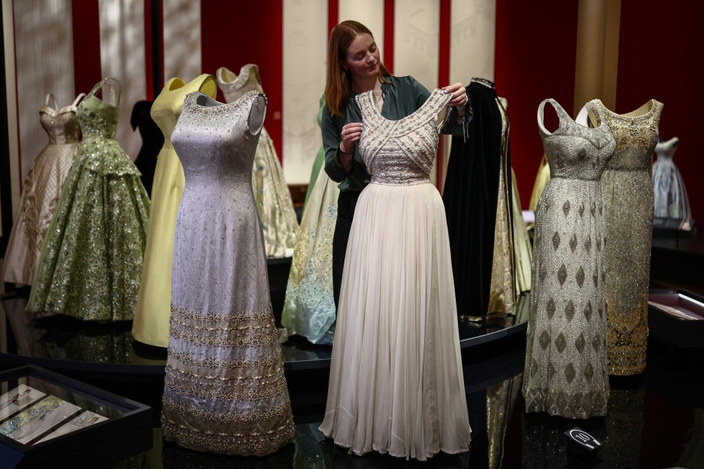 Photo by HENRY NICHOLLS / AFP  A member of the Royal Collection Trust staff poses with an evening dress (C), circa 1960, during a media preview of the exhibition 'Queen Elizabeth II: Her Life in Style' at the King’s Gallery in Buckingham Palace in London on April 9, 2026.