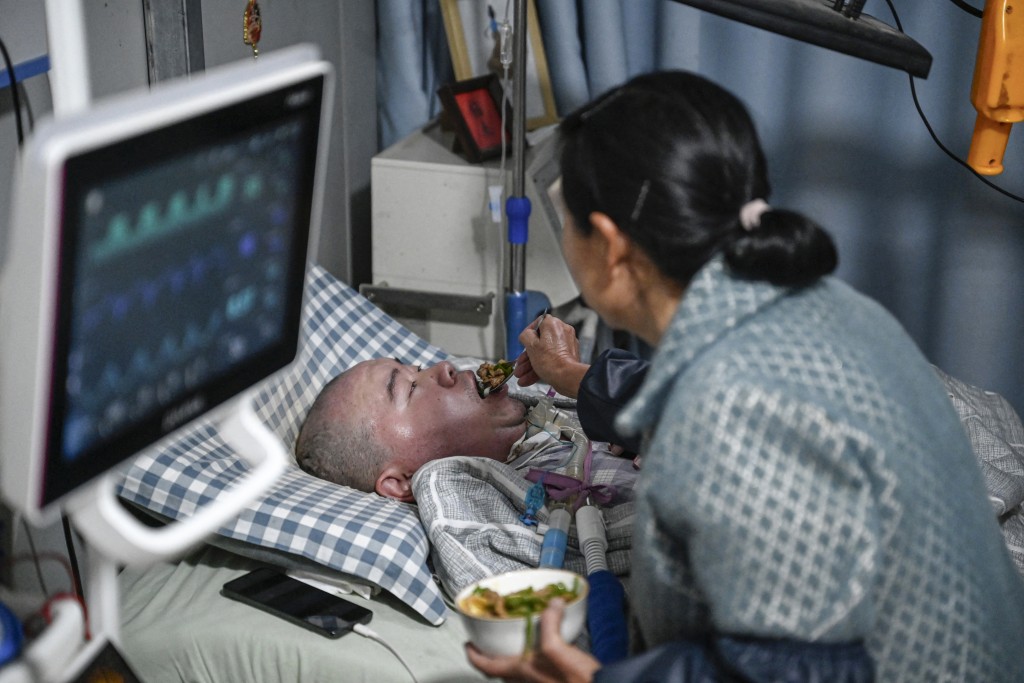 Photo by JADE GAO / AFP  This photo taken on December 16, 2025 shows Wu Dimei feeding her son Li Xia, who has Duchenne muscular dystrophy at their house in southwestern China's Chongqing municipality.