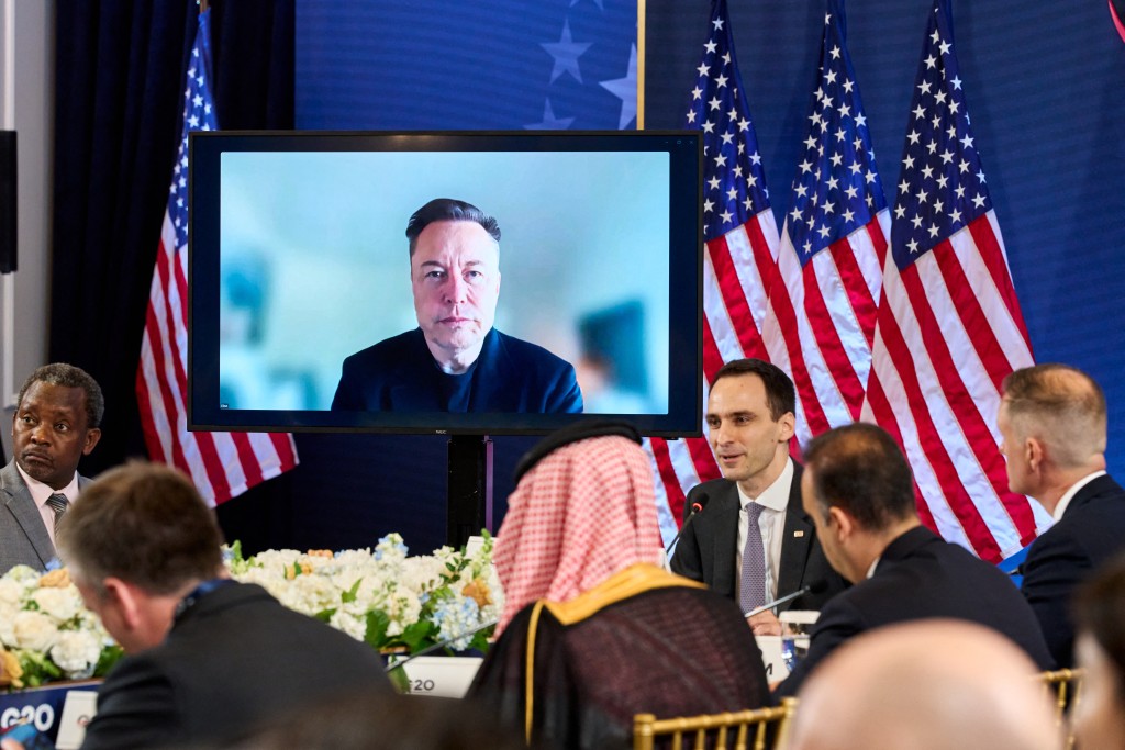 Elon Musk is seen on a screen as he virtually attends the G20 Innovation Ministerial summit next to the White House Office of Science and Technology Policy Director Michael Kratsios in Chapel Hill, North Carolina, on September 1, 2026. (Photo by Matt RAMEY / AFP)