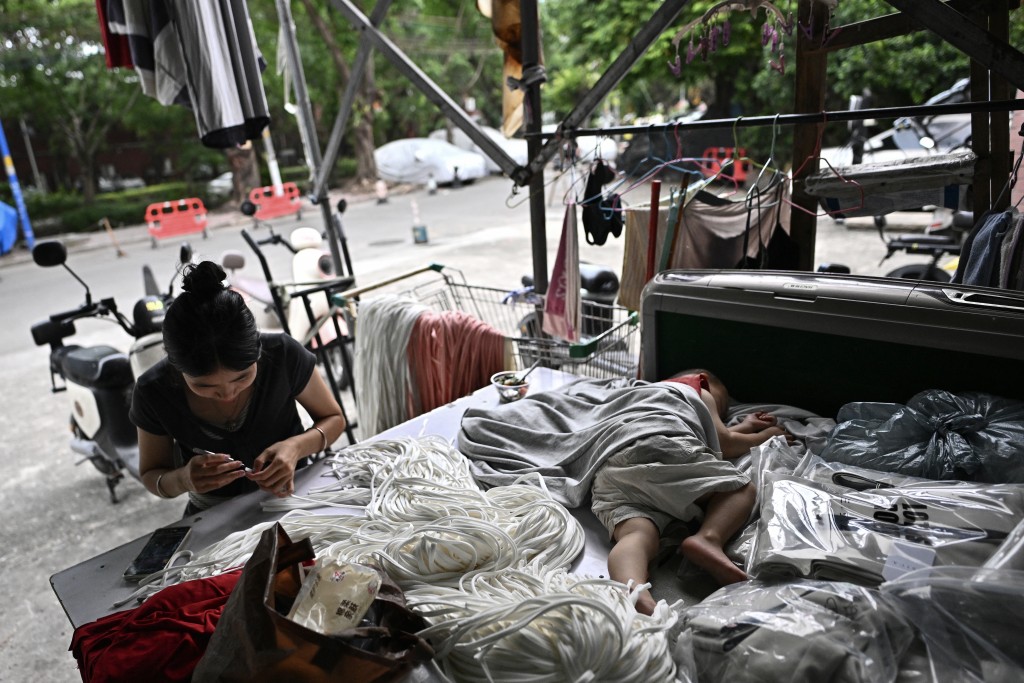 Photo by PEDRO PARDO / AFP  This picture taken on April 27, 2026 shows a woman working as her son sleeps next to her at a clothing sewing workshop in an urban village in Guangzhou, in southern China's Guangdong province.