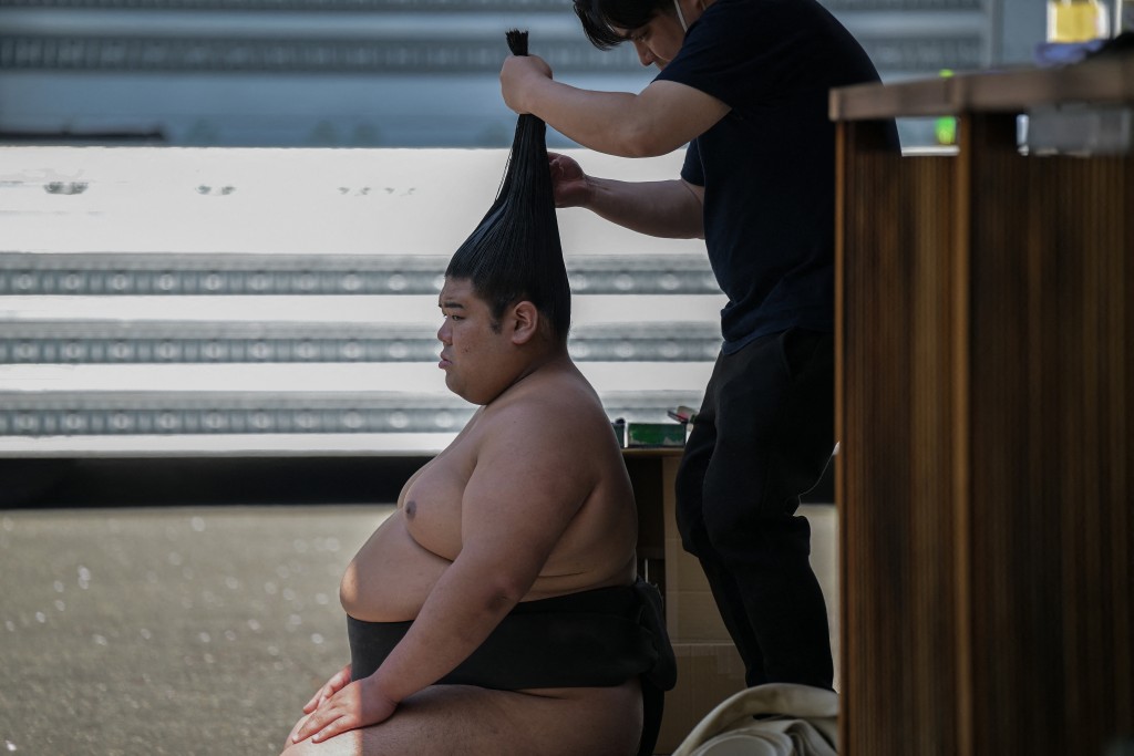 Photo by RICHARD A. BROOKS / AFP  A sumo wrestler has his hair prepared into a "top knot" before taking part in the annual "honozumo", a ceremonial one-day sumo exhibition for spectators held at Yasukuni Shrine in Tokyo on April 15, 2024.