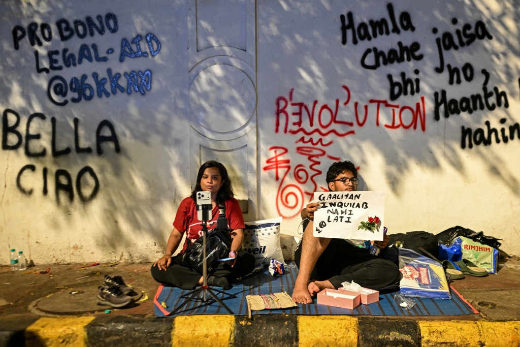 Photo by ARUN SANKAR / AFP This photograph taken on July 24, 2026 shows a Cockroach Janta Party (CJP) supporter recording a video while sitting beside wall graffiti against mainstream television media during a mass youth-led protest over exam irregularities at Jantar Mantar in New Delhi.