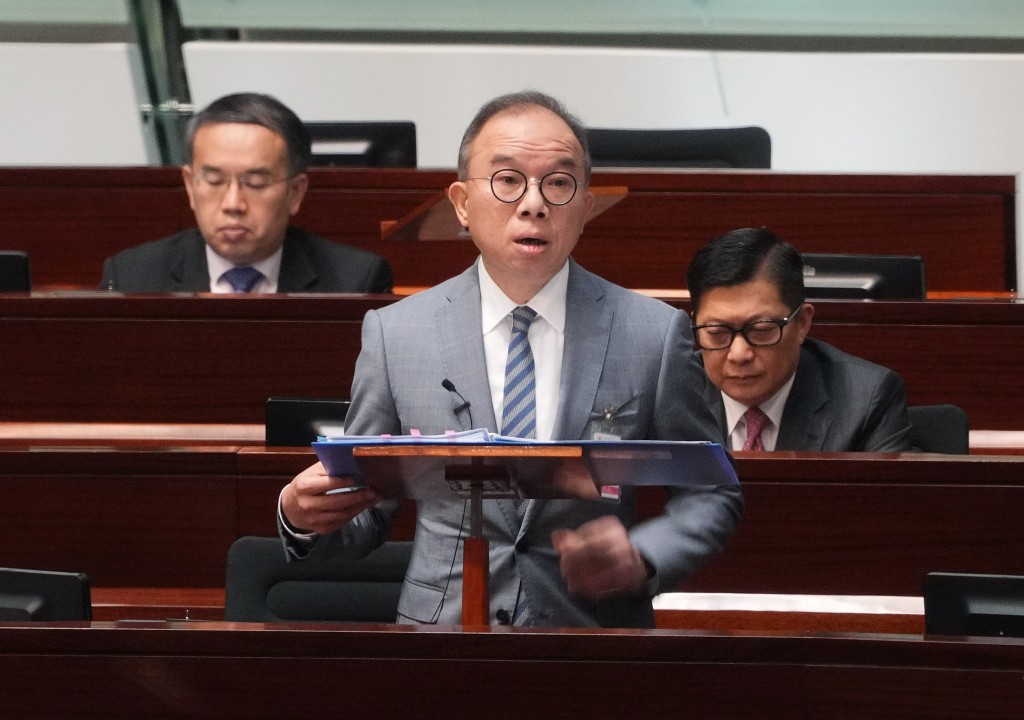 A man in a gray suit and glasses is speaking at a podium with a document in a legislative setting, while two other men in suits are seated behind him, with the background indicating a formal parliamentary environment discussing societal harmony and societal sensitivities related to the Same-Sex Partnerships Bill.