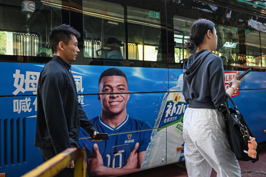 Photo by JADE GAO / AFP  People walk past a bus featuring French football star Kylian Mbappe for Dongpeng Beverage Group's "Bushuila" electrolyte drink on a street in Shanghai on October 27, 2026.