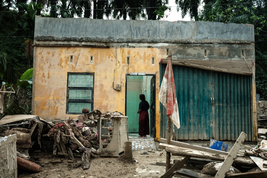 Photo by YASUYOSHI CHIBA / AFP  A woman stands inside a damaged house in Babo, Aceh Tamiang, Northern Sumatra, on December 15, 2025, following a flash flood.