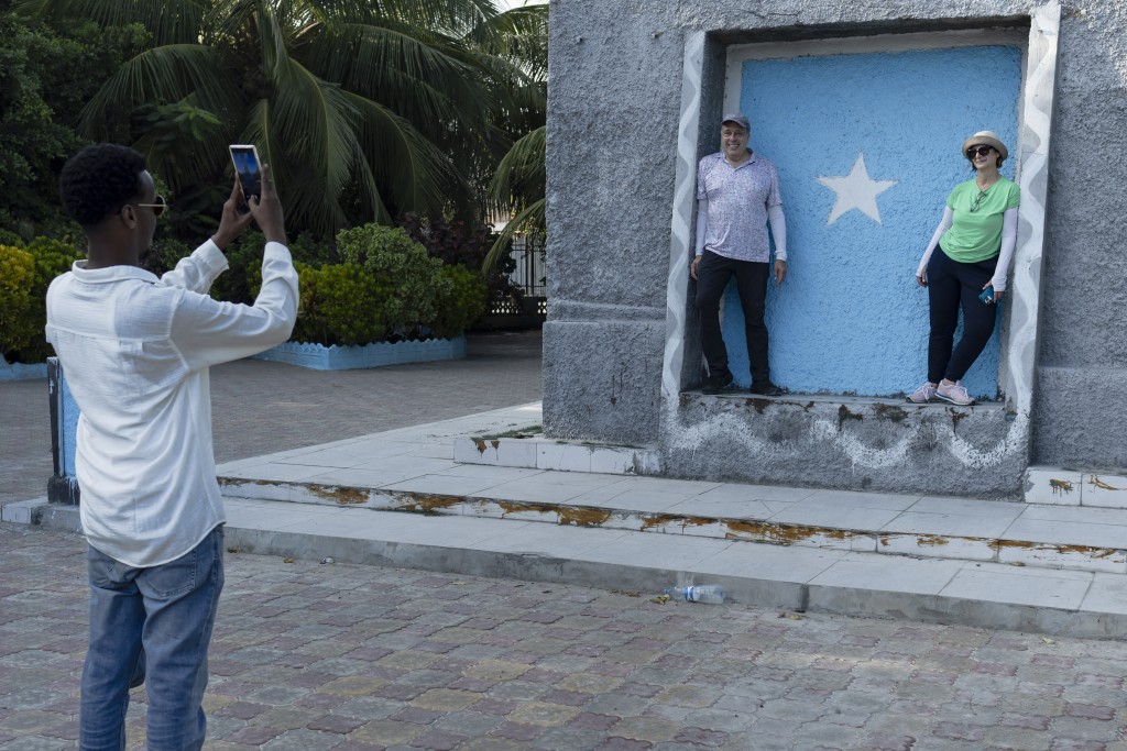 Photo by TONY KARUMBA / AFP  US tourists Richard (C) and Sheryl (R) pose for a photo taken by their tour guide at the Tomb of the Unknown Soldier monument during a guided tour of tourist attractions in Mogadishu on November 10, 2025.