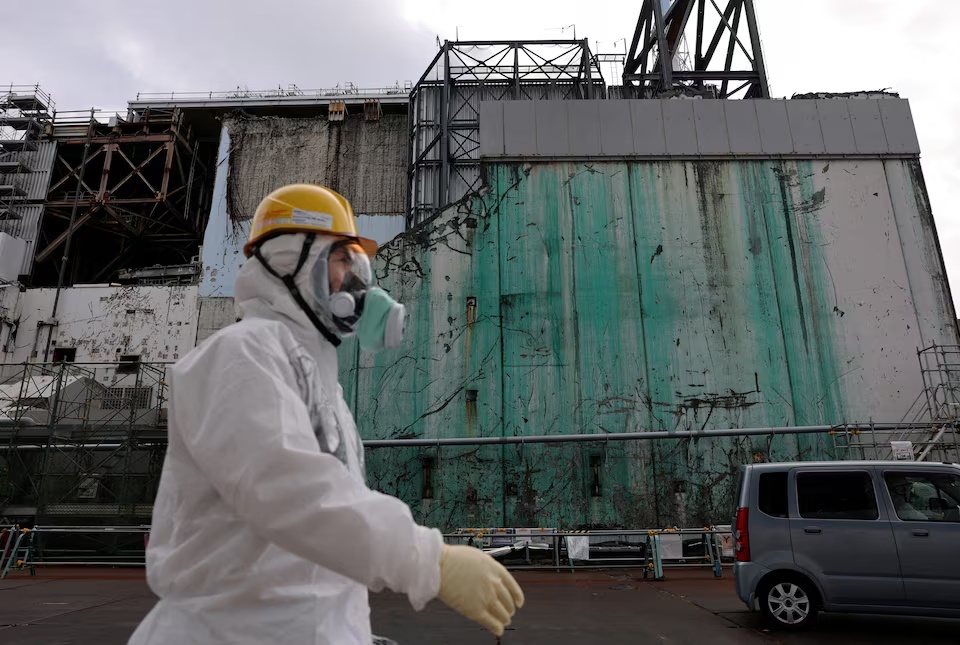 A worker wearing a protective suit and mask walks past the wall of the No. 3 reactor building, green from when an agent was sprayed on it to prevent the spread of radioactive material, and with scratches and scrape marks from the tsunami, at the tsunami-crippled Fukushima Daiichi nuclear power plant in Okuma town, Fukushima prefecture, Japan, January 22, 2026.  REUTERS/Issei Kato/File Photo