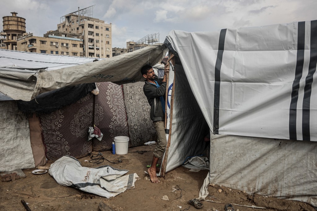 Photo by OMAR AL-QATTAA / AFP.  A Palestinian man fixes his makeshift shelter as the first winter rains fall on a displacement camp in Gaza City on November 14, 2025.