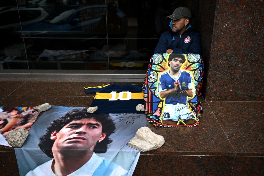 A supporter of Argentine football legend Diego Maradona sits beside portraits and memorabilia honoring Maradona outside the San Isidro courthouse during the final hearing of defense closing arguments in the trial over Maradona's death, in San Isidro, Buenos Aires Province, Argentina, on October 21, 2026. (Photo by Luis ROBAYO / AFP)