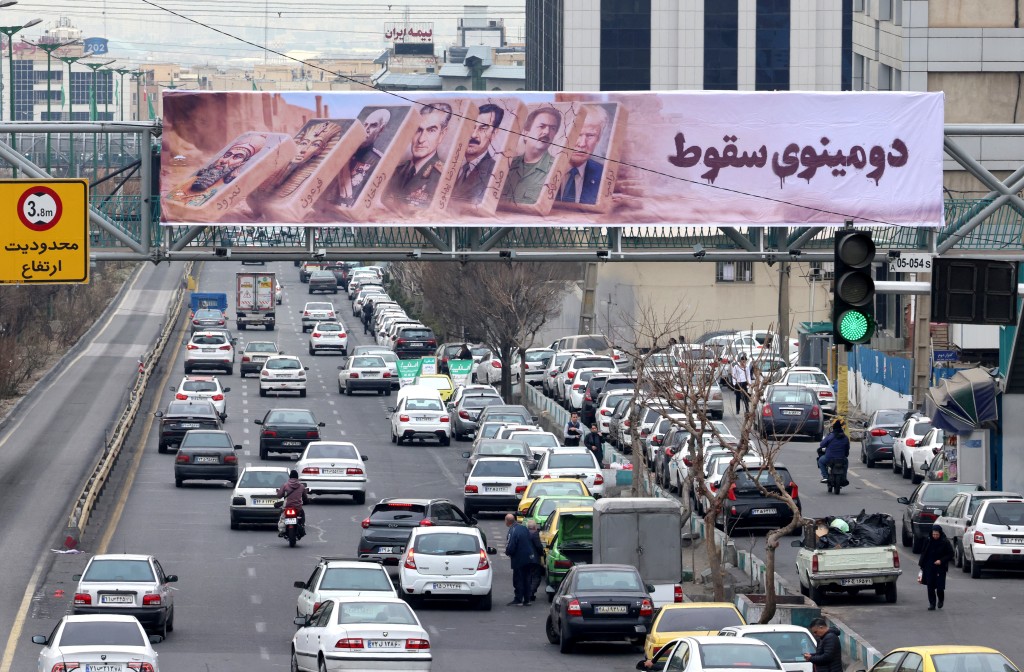 Photo by ATTA KENARE / AFP  Traffic rolls along a main throughfare under a banner with images of past and present leaders that reads in Farsi, "Domino fall", as daily life returns to the streets following nationwide protests, in the Iranian capital Tehran on January 19, 2026.