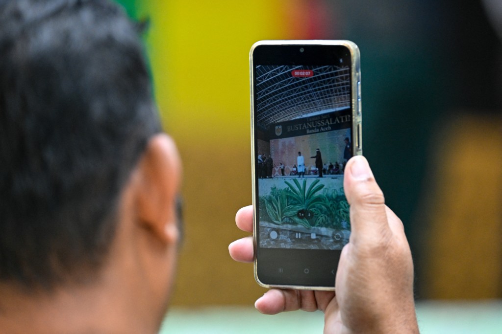 Photo by CHAIDEER MAHYUDDIN / AFP  / A man records a video as a man, part of an unmarried couple seen kissing on a Tiktok livestream, is caned as punishment under sharia law in Banda Aceh on July 2, 2026.