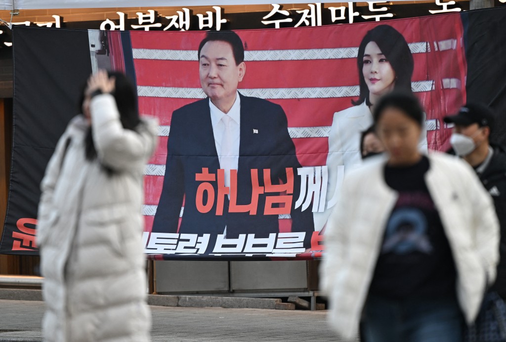 Pedestrians walk past a banner showing a picture of South Korea's former impeached president Yoon Suk Yeol and his wife Kim Keon Hee displayed by Yoon's supporters near the Seoul Central District Court in Seoul on January 9, 2026, as Yoon's final criminal trial on insurrection charges is held in the court. (Photo by Jung Yeon-je / AFP)