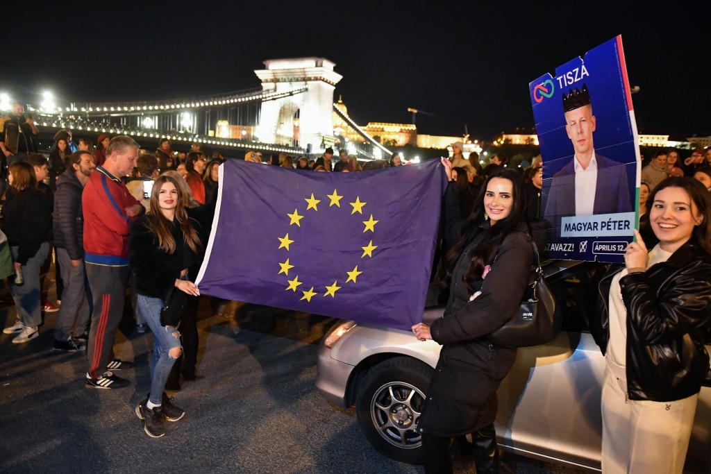 Photo by FERENC ISZA / AFP Participants display an election campaign poster of Hungary's prime minister-elect, Peter Magyar and a flag of the European Union during a spontaneous gathering to celebrate the victory of the conservative Tisza party near the Szechenyi Chain Bridge in Budapest late on April 13, 2026, one day after Hungary's general elections.
