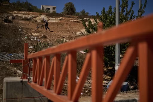 A Palestinian man pushes a wheelbarrow past a closed gate set up by Israeli authorities that blocks an entrance to the West Bank village of Sinjil, Tuesday, Sept. 30, 2025. (AP Photo/Leo Correa)