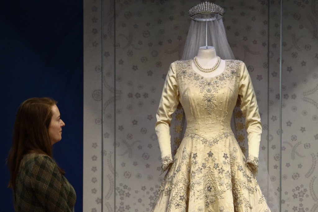 Photo by HENRY NICHOLLS / AFP  A member of the Royal Collection Trust staff poses next to 'Princess Elizabeth's Wedding Dress, Veil and Train' from 1947, during a media preview of the exhibition 'Queen Elizabeth II: Her Life in Style' at the King’s Gallery in Buckingham Palace in London on April 9, 2026.