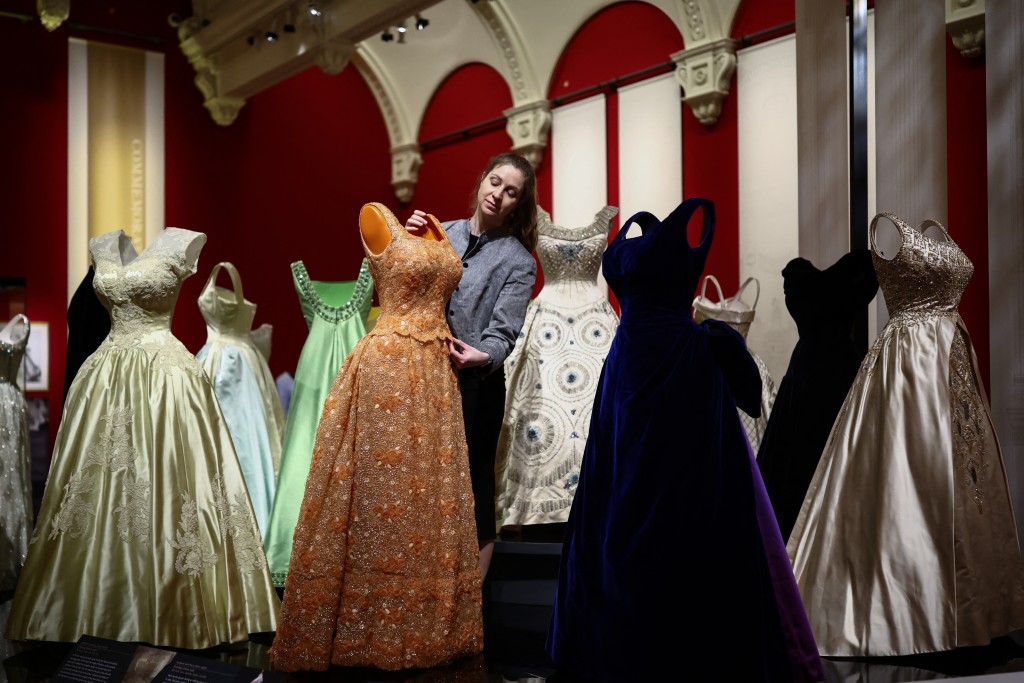 Photo by HENRY NICHOLLS / AFP  A member of the Royal Collection Trust staff poses with a display of evening gowns, during a media preview of the exhibition 'Queen Elizabeth II: Her Life in Style' at the King’s Gallery in Buckingham Palace in London on April 9, 2026.