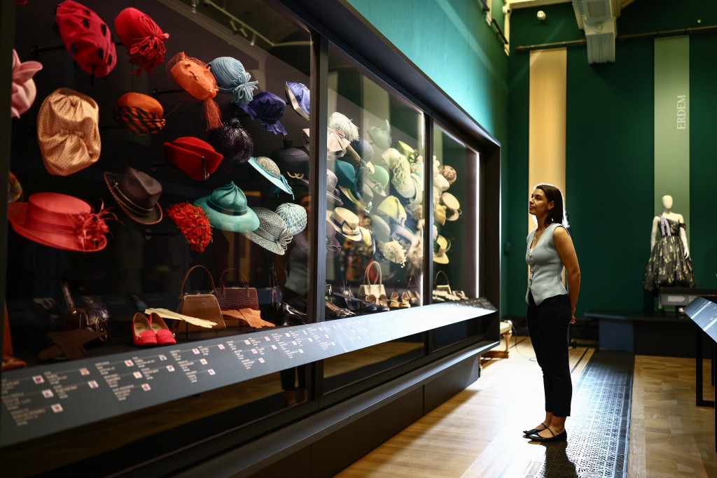 Photo by HENRY NICHOLLS / AFP  A member of the Royal Collection Trust staff poses next to a display of various hats, during a media preview of the exhibition 'Queen Elizabeth II: Her Life in Style' at the King’s Gallery in Buckingham Palace in London on April 9, 2026.