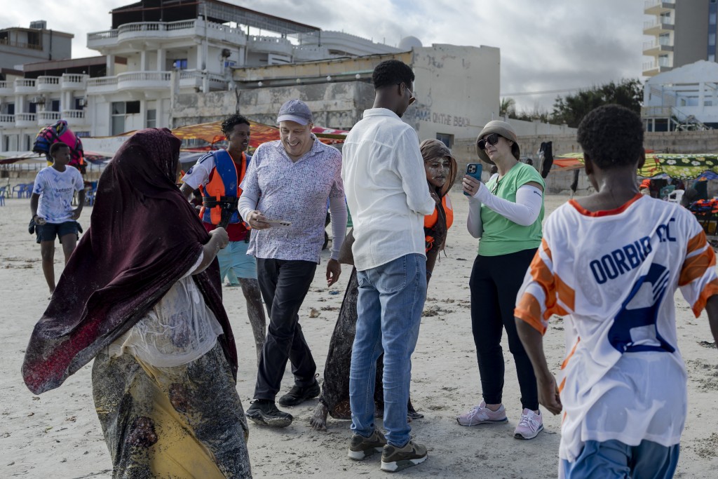 Photo by TONY KARUMBA / AFP  US tourists Richard (4th L) and Sheryl (2nd R) interact with local youth on their walk on Lido beach during a guided tour of tourist attractions in Mogadishu on November 10, 2025.