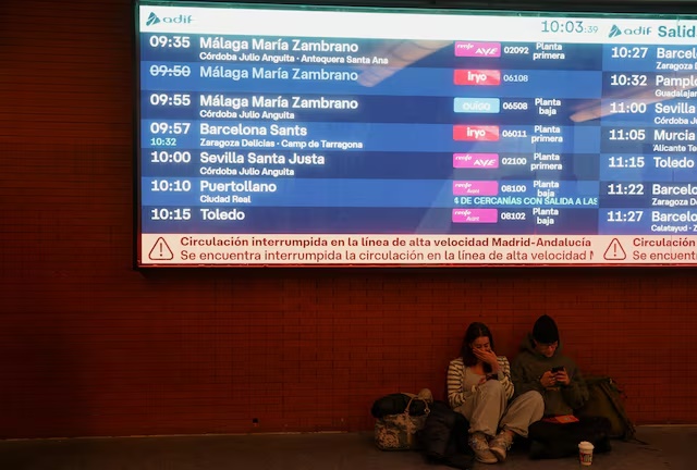 Two people sit below an information screen with a message stating that the high-speed train line between Madrid and Andalucia has been interrupted following the collision of two high-speed trains between the towns of Mora and Orgaz, at the Atocha train station in Madrid, Spain, October 28, 2026. REUTERS/Susana Vera