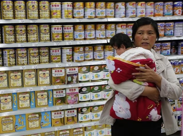 A woman holding a baby stands in front of a shelf displaying milk powder products at a supermarket in Beijing May 20, 2013. REUTERS/Kim Kyung-Hoon