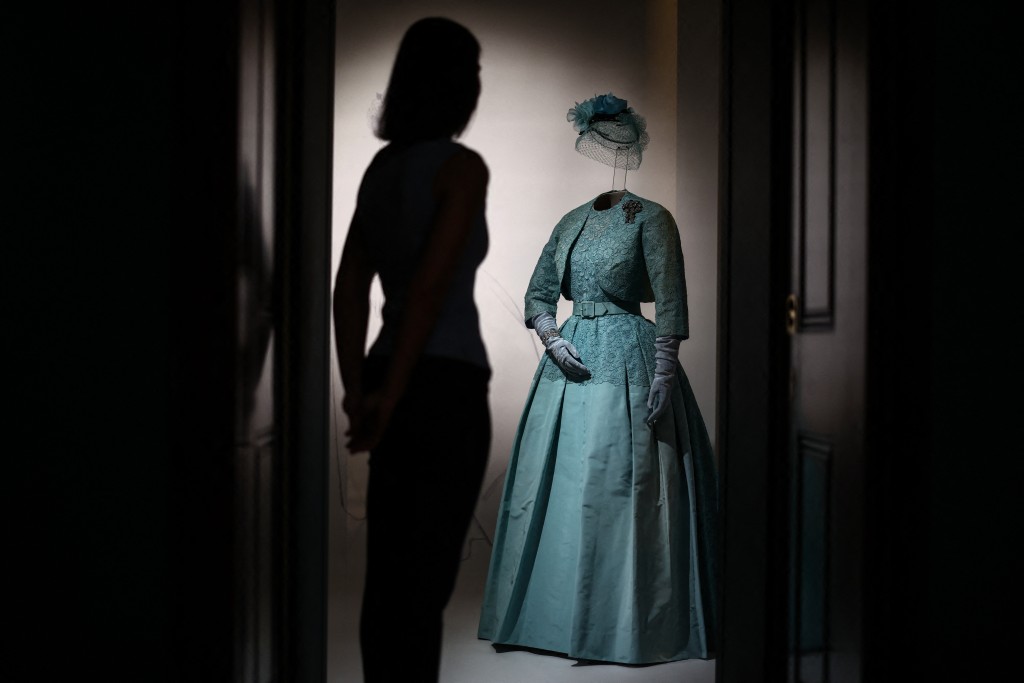 Photo by HENRY NICHOLLS / AFP  A member of the Royal Collection Trust staff (L) is silhouetted next to an ensemble worn for Princess Margaret's wedding in 1960, during a media preview of the exhibition 'Queen Elizabeth II: Her Life in Style' at the King’s Gallery in Buckingham Palace in London on April 9, 2026.