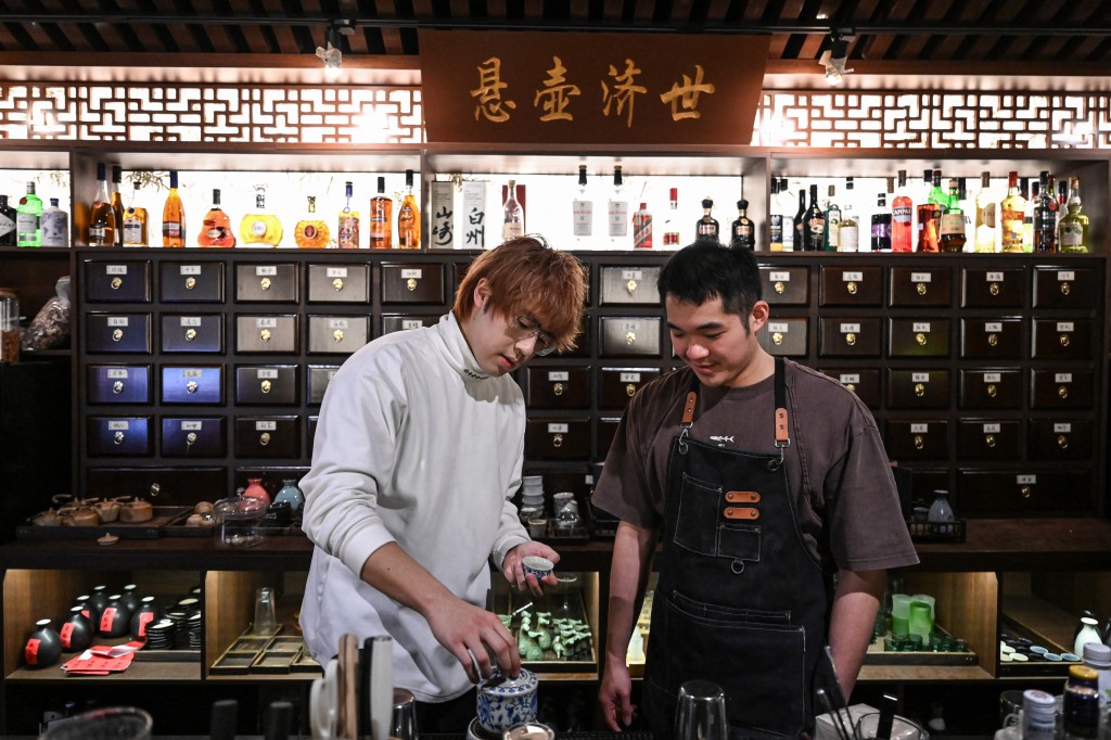 Photo by JADE GAO / AFP  / The photo taken on February 3, 2026 shows bar owner Wu Siyuan (L) talking with a bartender in front of a plaque with the motto of Chinese medicine “Practice medicine to help the world” at a traditional Chinese medicine-themed cocktail bar in Shanghai.