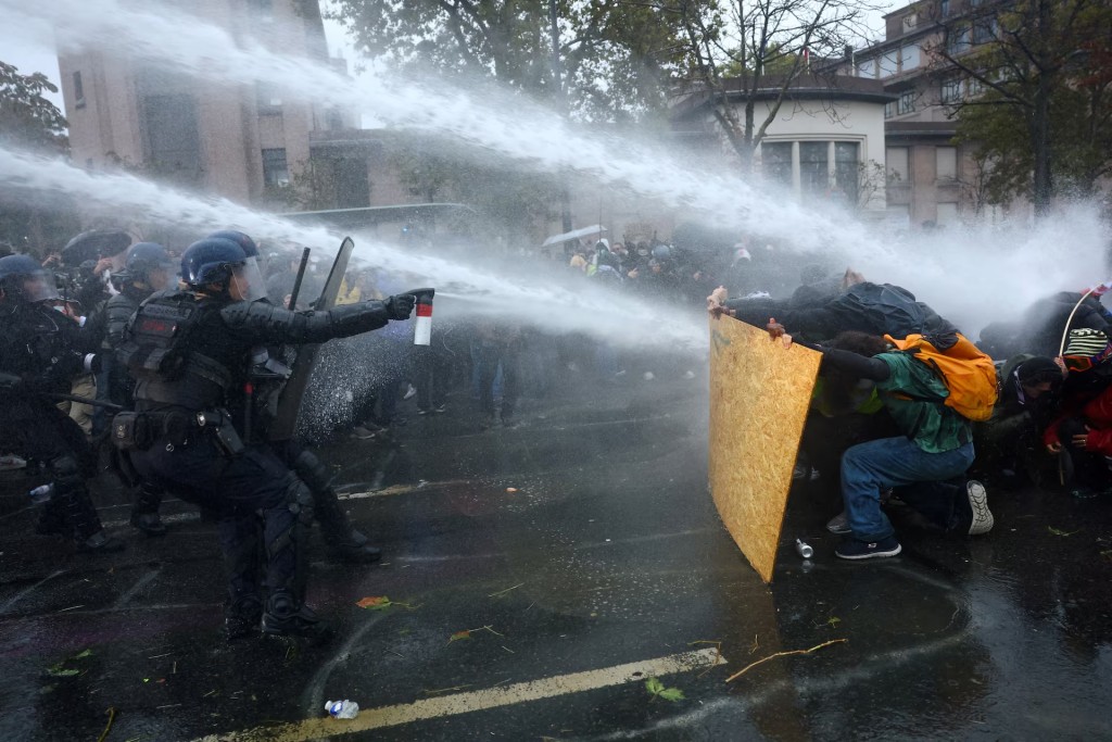 Protesters take cover behind a plywood board as French riot police and gendarmes use a water canon amid clashes during an authorised demonstration in Paris, October 24, 2026. REUTERS/Hannah McKay