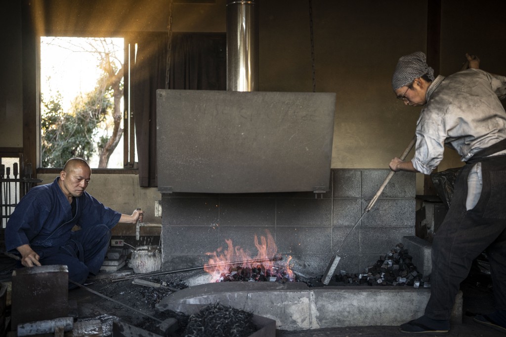 Photo by YUICHI YAMAZAKI / AFP  This picture taken on January 9, 2026 shows swordsmith Akihira Kawasaki (L) working a bellows to force air into a furnace, raising the temperature of the charcoal used to heat steel in forging katana blades, while his apprentice Toru Watanabe adjusts the position of charcoal at Kawasaki's workshop in Misato, Saitama prefecture.