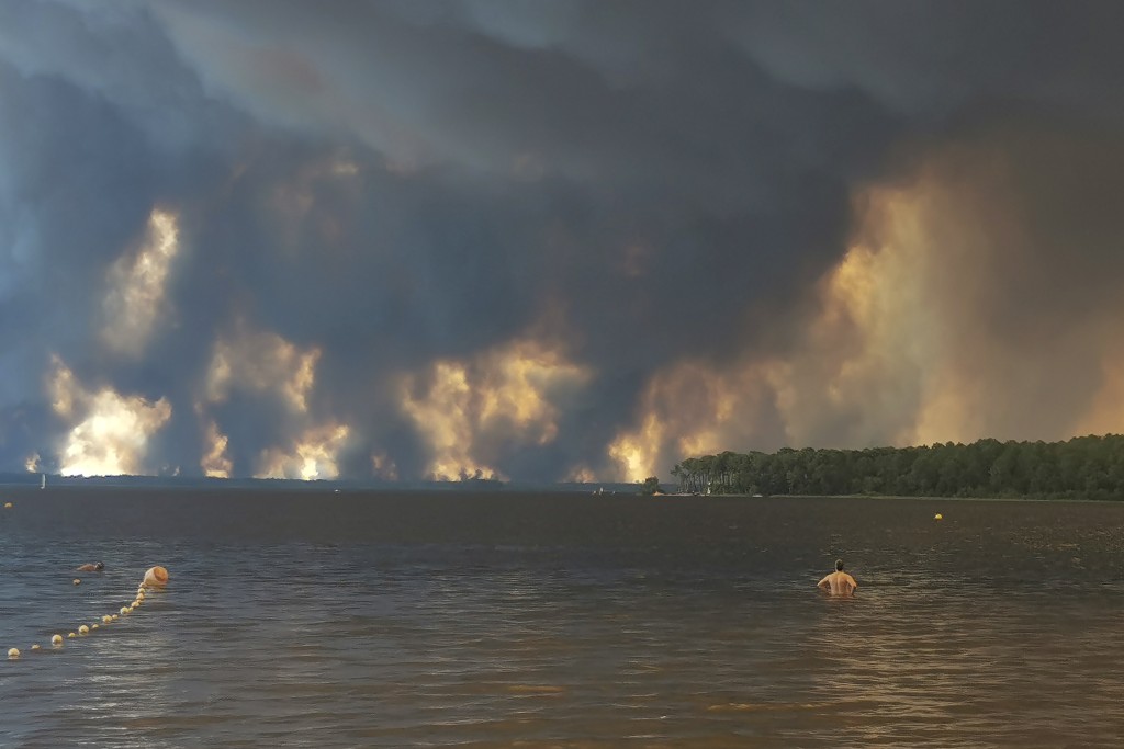 Photo by MAXIMILIEN LAMY / AFP  / A beach goer stands in lake Lacanau, also called Etang de Lacanau (Lacanau Pond) from the beach of Moutchic in Lacanau, southwestern France on July 24, 2026, as clouds of smoke rise into the sky from a wildfire.