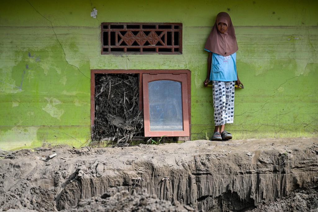 Photo by CHAIDEER MAHYUDDIN / AFP  A child stands on hardened mud beside the window of a residence damaged by flash floods in Meurah Dua, Aceh province's Pidie Jaya district on February 21, 2026, after devastating floods and landslides struck Indonesia's Sumatra late last year.