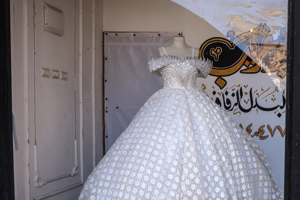 Photo by BASHAR TALEB / AFP A wedding gown stands behind the shattered vitrine of workshop where dresses are created including evening and wedding gowns despite limited resources and old dresses are recycled, in the city of Khan Yunis in the southern Gaza Strip on June 13, 2026.