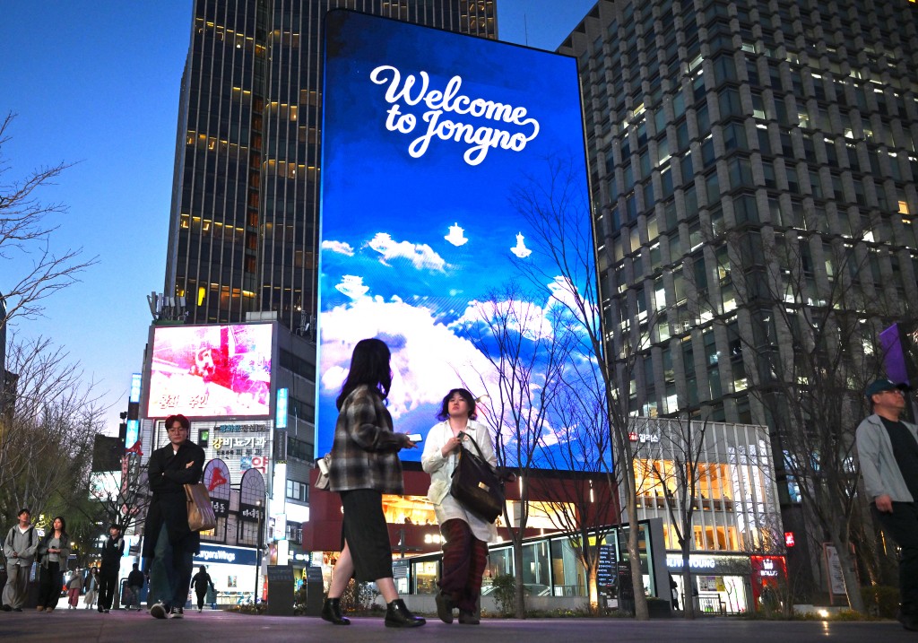 Photo by JUNG YEON-JE / AFP. People walk in front of digital billboards at Gwanghwamun Square in Seoul on April 1, 2026.