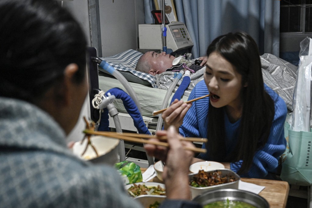 Photo by JADE GAO / AFP  This photo taken on December 16, 2025 shows Li Xia (back), who has Duchenne muscular dystrophy, sharing a meal with his family at their house in southwestern China's Chongqing municipality.