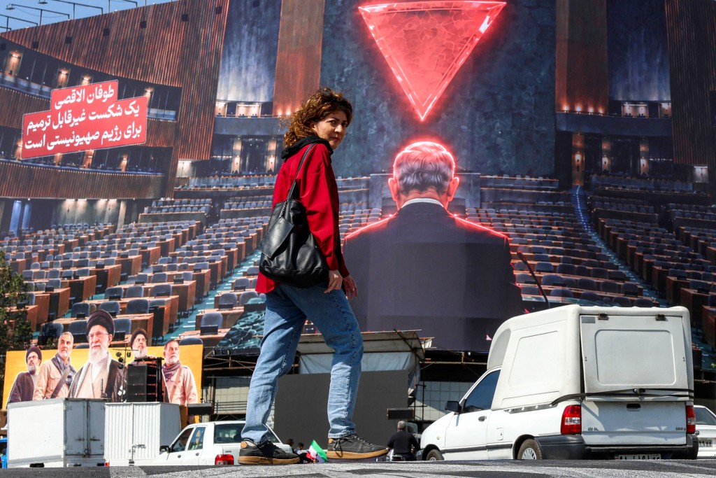 Photo by - / AFP A woman walks through traffic while crossing Enghelab square in central Tehran October 12, 2026, as behind a large anti-Israeli billboard is displayed.