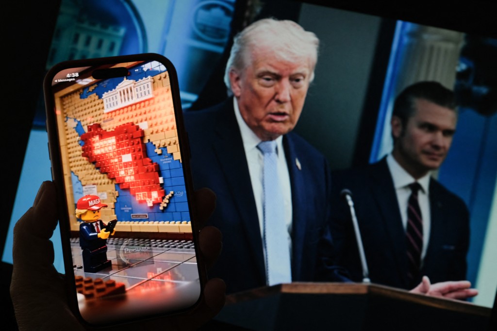 Photo by CHRIS DELMAS / AFP  This illustration photo created in Los Angeles on April 9, 2026 shows a Lego-style AI-generated war-themed video playing on a smartphone screen in front of a photo of US President Donald Trump and US Secretary of Defense Pete Hegseth.