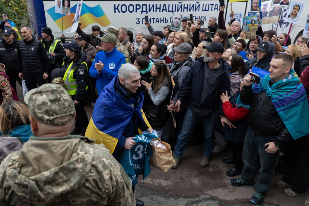 Photo by SERHII OKUNEV / AFP  A released Ukrainian prisoner of war (POW) wrapped in the national flag walks past people holding portraits of their missing or captured relatives, following a prisoners exchange in an undisclosed location in Ukraine on October 23, 2026, amid the Russian invasion of Ukraine.