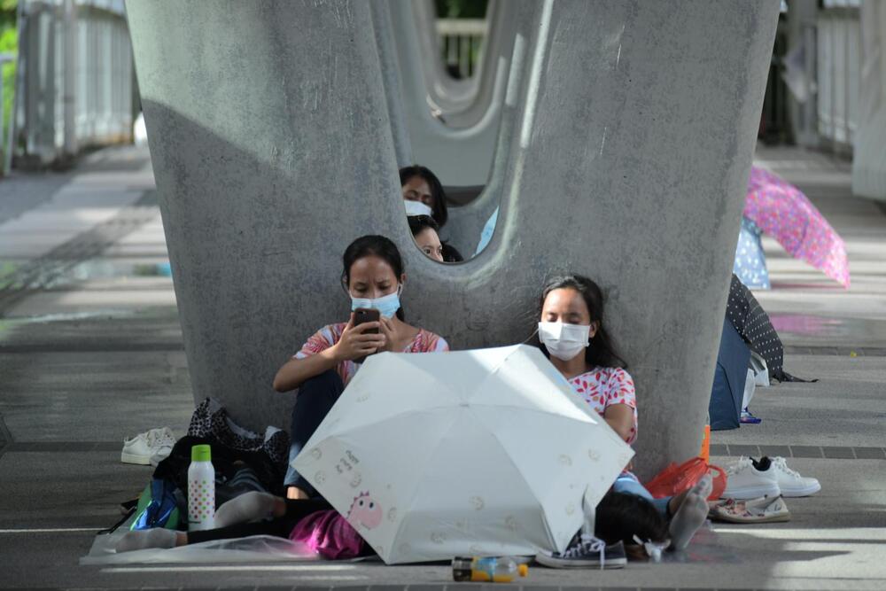 A group of women, wearing face masks, are sitting on the ground beneath a large structure with their personal belongings and umbrellas, possibly reflecting a moment of waiting or mourning related to a tragedy involving a Filipino domestic helper.