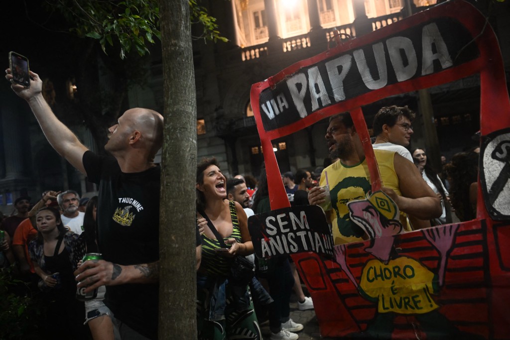 A man stands behind a drawing of a bus to Papuda, name of a Brazilian high security prison, during a street party to celebrate the Brazilian Supreme Court's decision to sentence former President Jair Bolsonaro in downtown Rio de Janeiro, Brazil on September 12, 2025. Brazil's Supreme Court on September 11 sentenced firebrand ex-president Jair Bolsonaro to 27 years in prison for coup plotting at the end of a landmark trial that divided the nation and drew US fury. (Photo by MAURO PIMENTEL / AFP)