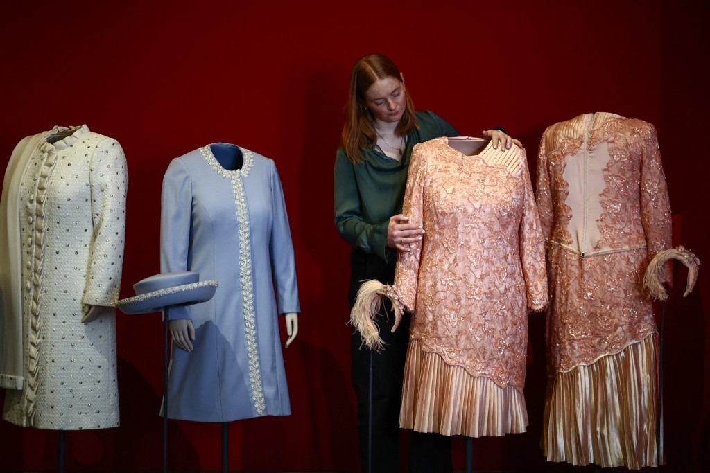 Photo by HENRY NICHOLLS / AFP  A member of the Royal Collection Trust staff poses with 'Two Dresses and Headpieces', from the 2012 London Olympics opening ceremony, during a media preview of the exhibition 'Queen Elizabeth II: Her Life in Style' at the King’s Gallery in Buckingham Palace in London on April 9, 2026.