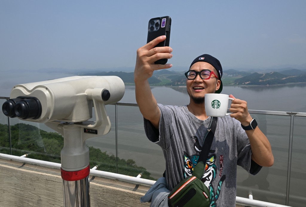 Photo by JUNG YEON-JE / AFP  A visitor holding a Starbucks cup takes a selfie as he looks at the North Korean side from a South Korean observation deck at Aegibong Peace Ecopark in Gimpo on July 1, 2026.