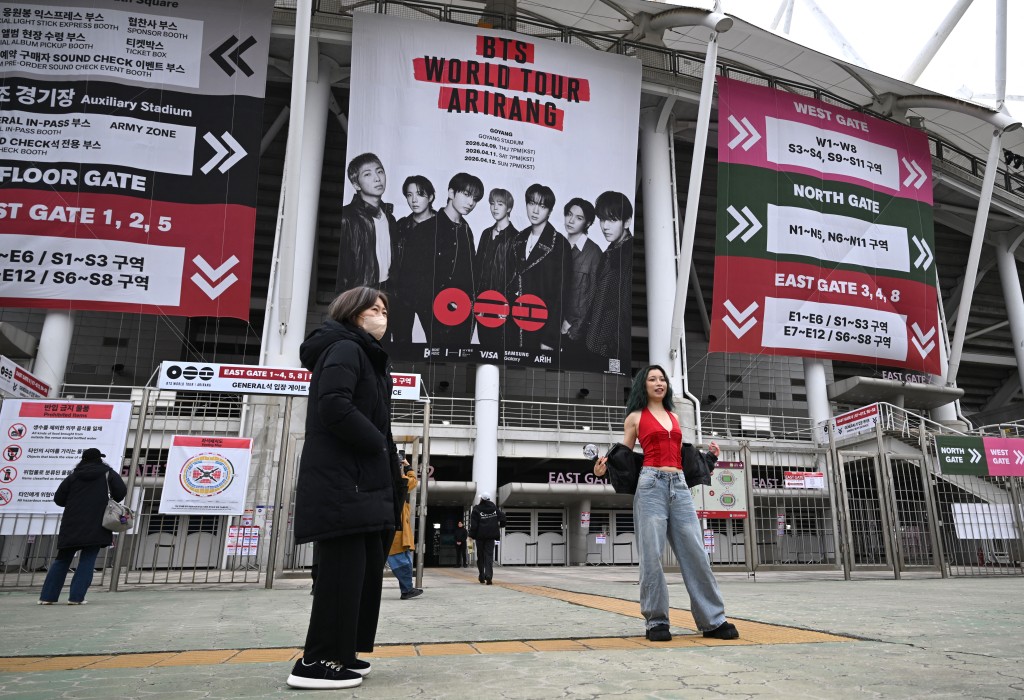 Photo by JUNG YEON-JE / AFP  A woman poses in front of a large banner featuring K-pop boy band BTS at a stadium where the group will perform in Goyang on April 9, 2026.