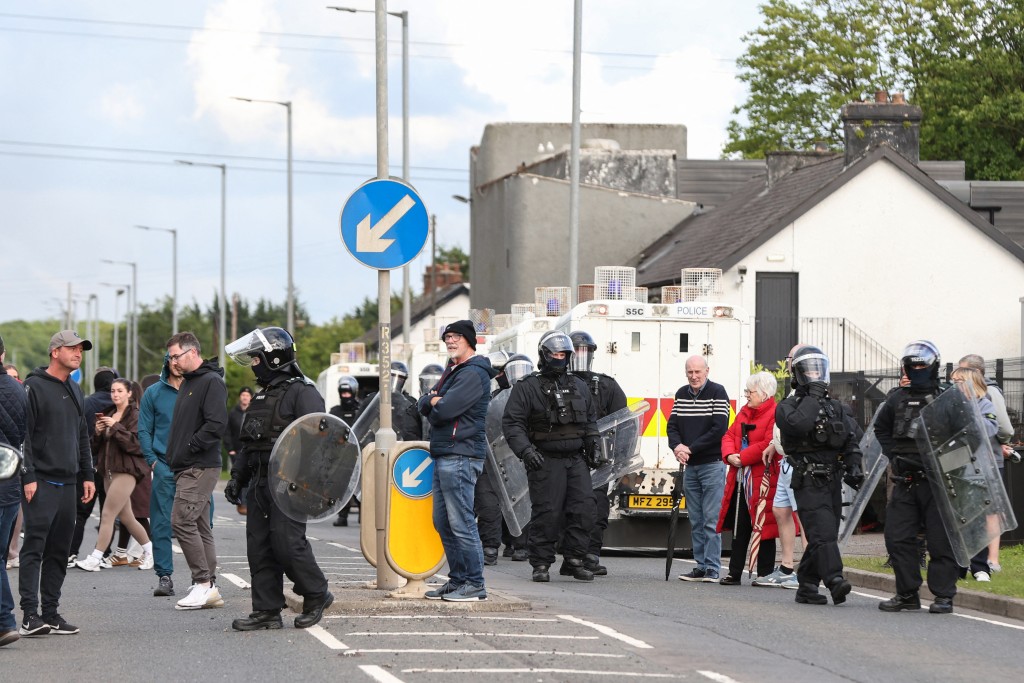 Photo by PAUL FAITH / AFP People stand near police in riot gear (R) as they seal off the road leading to a hotel previously believed to house migrants, in the town of Mallusk, Co Antrim, north of Belfast, Northern Ireland, on June 10, 2026.