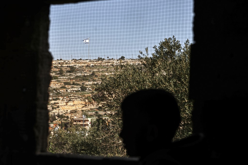 Photo by ZAIN JAAFAR / AFP  This picture taken from Umm Safa village, north of Ramallah in the occupied West Bank, shows a Palestinian youth silhouetted against a window with an Israeli flag fluttering on a hilltop overlooking the village on February 16, 2026.